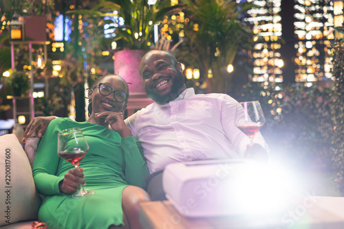 фотографія Joyous couple watches projected comedy during a rooftop restaurant date beneath glowing city lights, laughing at funny scenes together while drinking red wine during their night out