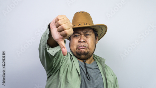 Картината върху платно Showing strong disapproval, one young Asian male wearing a brown cowboy hat extends a downward pointing thumb toward the viewer against some plain light background