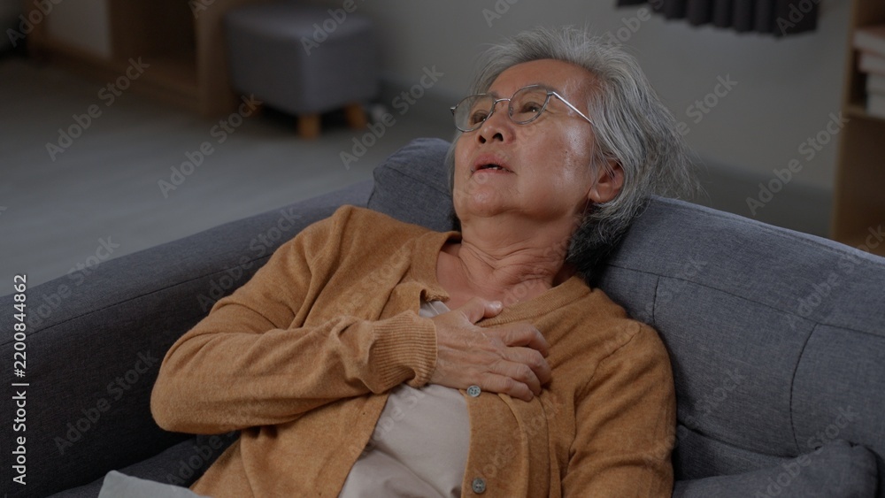 Fototapeta premium Senior woman reclines on a gray sofa with one hand on her chest, looking upward and breathing through visible discomfort while experiencing sudden upper body pain alone inside her own home.