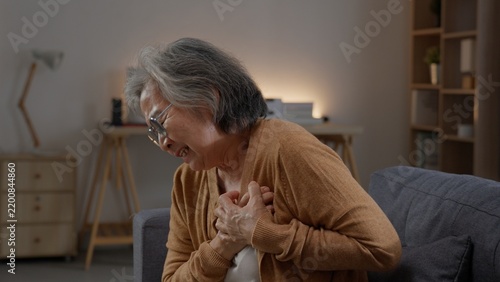 Obraz na płótnie Elderly woman bends forward on a sofa while pressing both hands to her chest, grimacing with intense discomfort during a sudden episode of upper body pain while staying alone at home today