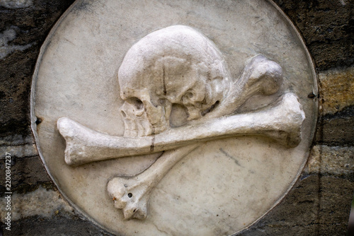 Fotografie Carved stone bas relief medallion featuring human skull and crossbones on old cemetery gravestone, representing mortality, memento mori symbol, and gothic historical masonry