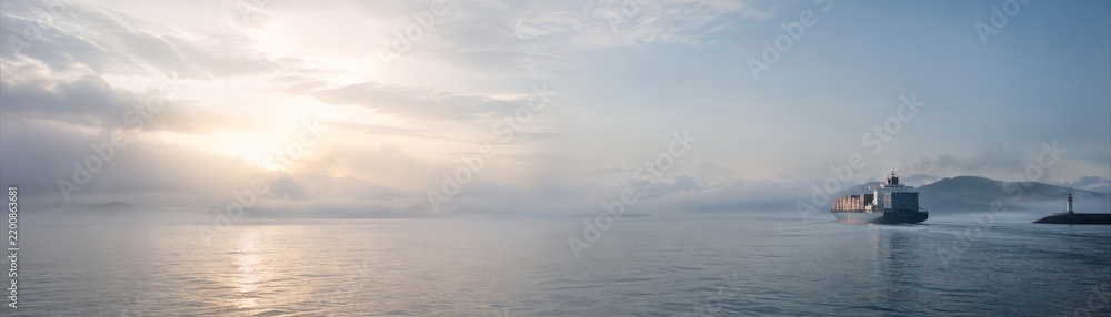 Obraz premium Panoramic view of cargo ship sailing through foggy coastal waters at golden sunrise over mountain horizon
