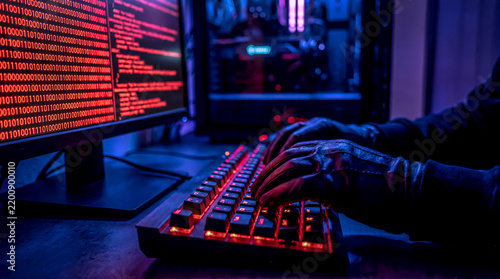 Close-up of hands typing on illuminated keyboard with red code on computer monitor screen, hacking and programming concept.