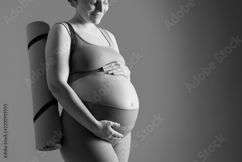 Sporty young pregnant woman with yoga mat on grey background, black-and-white...