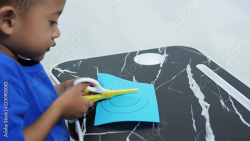 A handsome Asian preschool-aged boy is concentrating on cutting out a pattern from blue paper to practice his motor skills, using child-friendly yellow-and-white scissors.