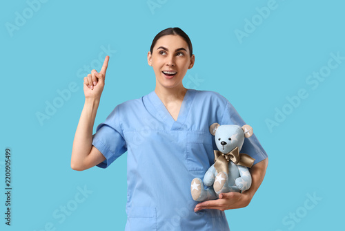 Young female pediatrician with teddy bear pointing at something on blue backg...