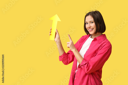 Beautiful young woman with paper arrow pointing at something on yellow backgr...