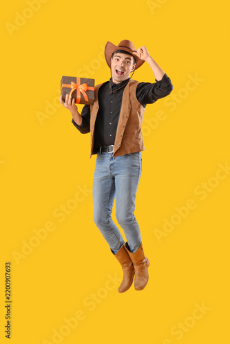 Young man dressed for Halloween as cowboy with gift jumping on yellow background