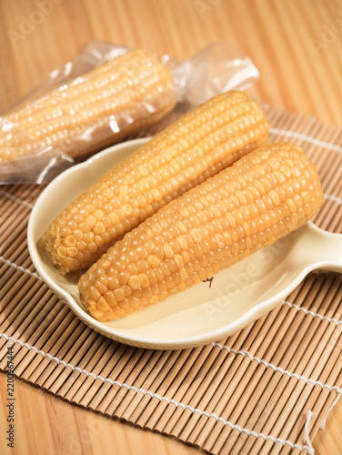 Obraz na płótnie Two Yellow Corn Cobs in Handled White Dish with Vacuum Packed Corn Background