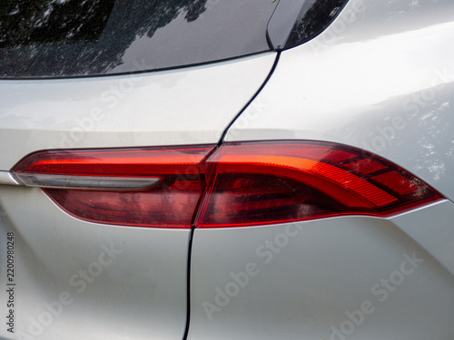 Close-up detail of a modern white car rear taillight, featuring sleek red LED lighting, aerodynamic bodywork, glossy painted surface, and reflections of trees and sky. Automotive exterior detail photo