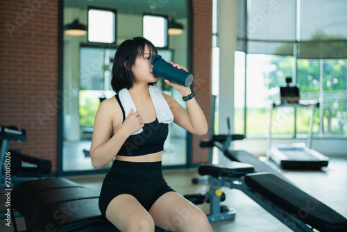 Young woman in black workout outfit drinking water from bottle sitting on bench in modern gym fitness studio