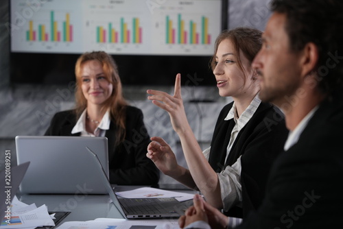 Принти на полотні Open hand lifted mid-argument, the young businesswoman presses her proposal while her ginger-haired colleague watches with amused approval at the dark table in a bright modern coworking