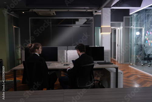 Fotografie Two colleagues seen from behind work side by side at monitors in a dark open plan office where warm lamps and glass partitions punctuate the late evening gloom in a bright modern coworking