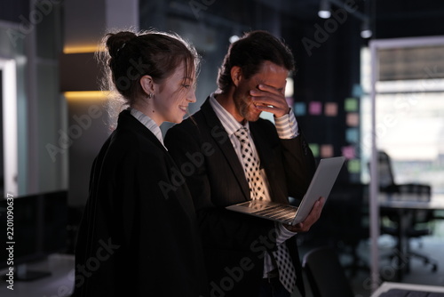 Фотография Full facepalm from the long haired colleague as he covers his eyes over the laptop and the young businesswoman laughs openly in the dark office in a bright modern coworking office