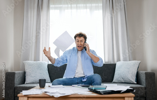 Angry man on phone throws overdue utility bills in despair. He faces severe financial pressure, rising energy expenses, and an economic crisis fueled by inflation.