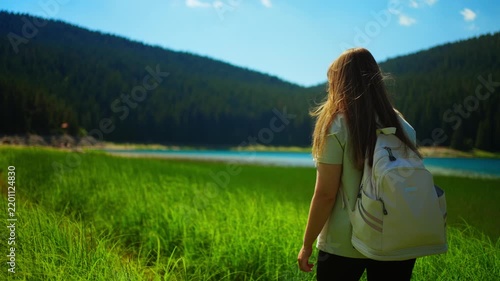 Young traveler with backpack walking through lush green meadow toward tranquil mountain lake surrounded by dense forest under bright summer sky