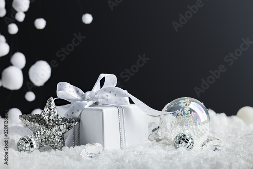 White Christmas gift with silver star and clear ornaments in snow on black background for festive New Year greeting banner