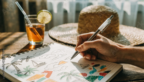 Person drawing in a sketchbook with a straw hat and iced tea on a table