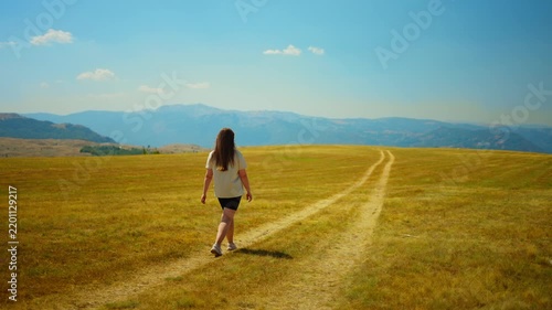 Solitary woman walking along a winding dirt path through a vast golden meadow under a clear blue sky with distant mountains on a bright summer day