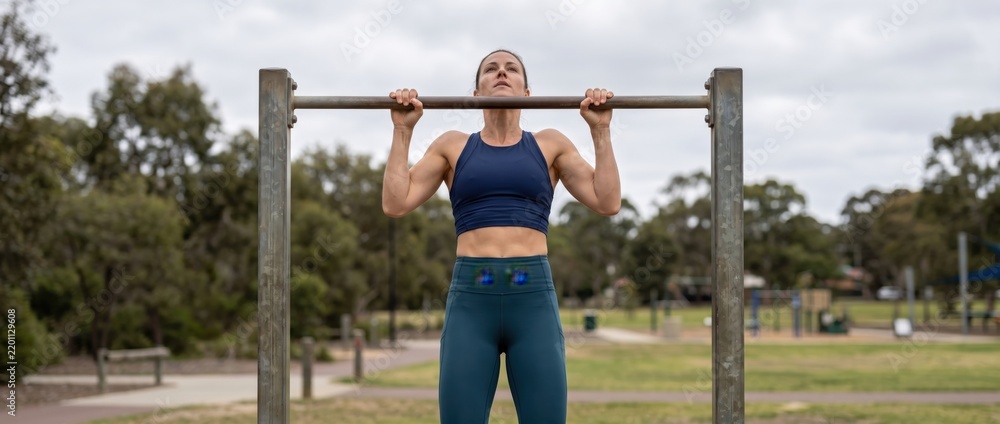 Fototapeta premium A fit woman performing pull-ups on a metal horizontal bar at an outdoor park, wearing a dark blue sports bra and teal leggings, with trees and a grassy field in the background