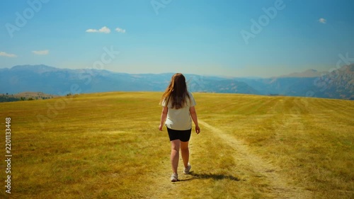 Young woman walking alone along a winding dirt path through a wide golden meadow toward distant hazy mountains under a clear blue summer sky