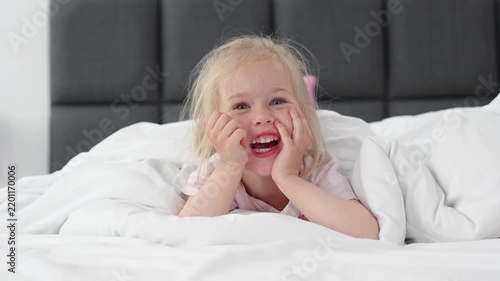 Caucasian blonde little girl child lying on a bed on white sheets, smiling and laughing at the camera