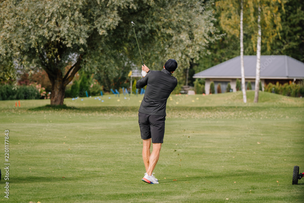 Fototapeta premium Golfer in dark clothing completing a full swing across manicured fairway, viewed from behind with trees and clubhouse in the background at an outdoor golf course