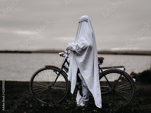 Obraz na plátně A person dressed as a white sheet ghost wearing sunglasses stands with a bicycle beside a lake