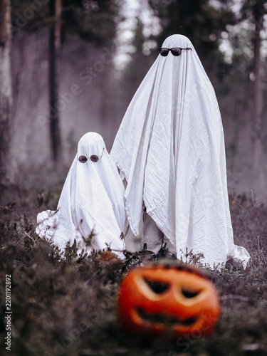Obraz na plátně Two people dressed as white sheet ghosts wearing sunglasses stand together in a forest with a Halloween pumpkin in the foreground
