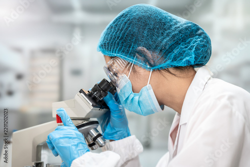 Female laboratory scientist in protective gear looks through a microscope while conducting research in a clean modern lab.