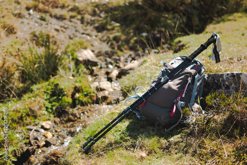 Hiking backpack and trekking poles leaning on a rock next to a flowing stream in a sunlit grassy mountain landscape, representing a break from an adventurous outdoor journey