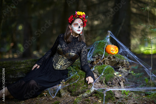 Halloween beauty portrait of young woman in the forest on a mossy stump with a halloween pumpkin.
