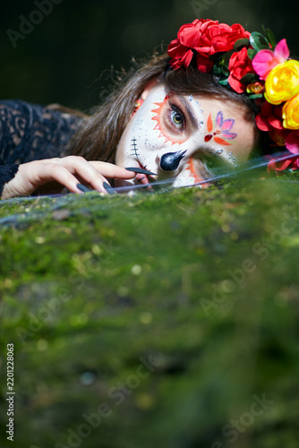 Halloween beauty portrait of young female with Santa Muerte makeup lies on tree trunk in the forest