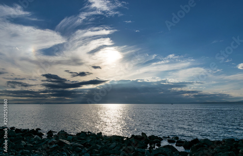 Sunlight over ocean horizon with clouds and rocky shoreline.