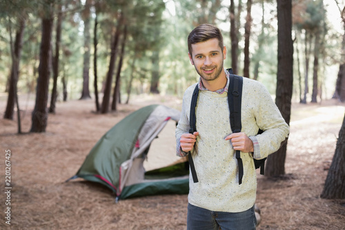 Adult male standing in pine forest campsite holding backpack straps near green tent wearing sweater