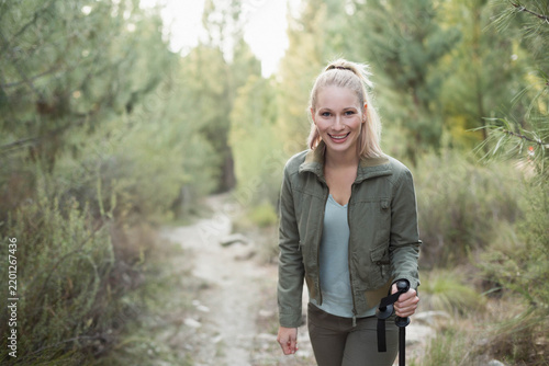 Female hiker standing on dirt trail in forest, holding trekking pole and wearing olive jacket