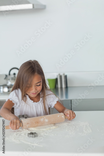 Asian girl child in blouse pressing and rolling dough on floured kitchen counter with wooden pin
