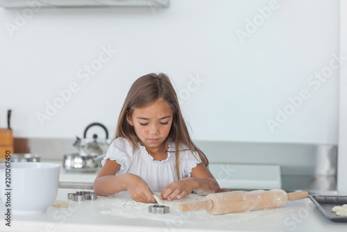 Child girl in white ruffled blouse shaping cookie dough at kitchen counter with wooden rolling pin