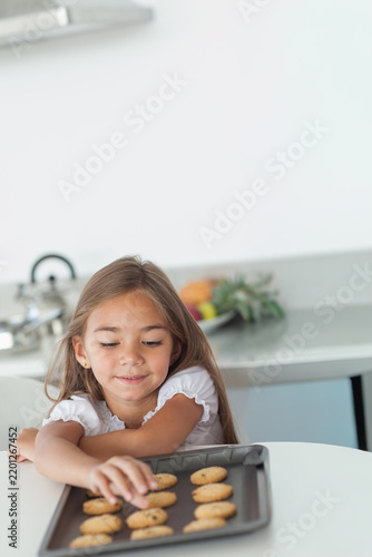 Child girl in white ruffled top sitting at kitchen table reaching for cookie on metal tray
