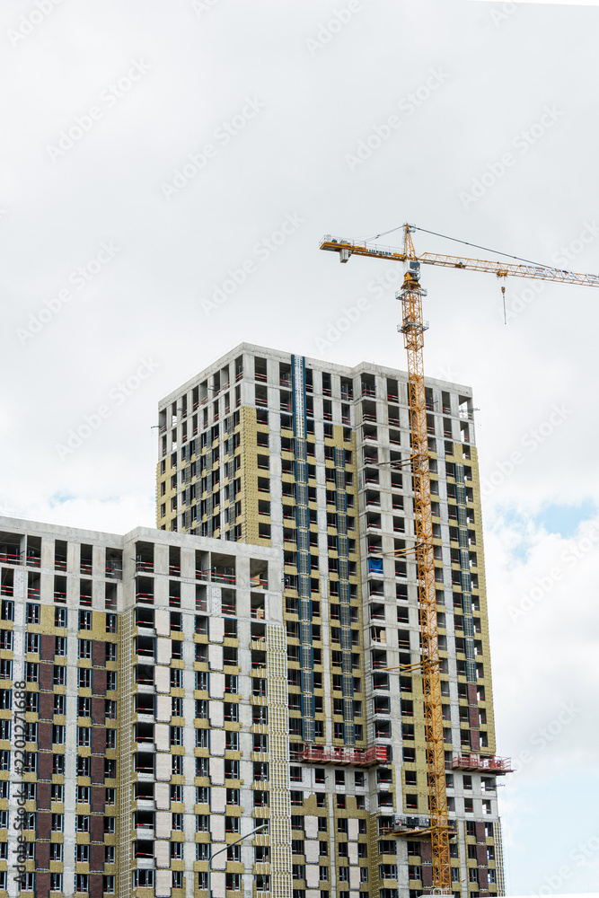 Fototapeta premium Residential skyscraper under construction with tower crane against cloudy sky. Urban development and real estate industry concept. Vertical format.