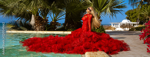 Stunning blonde model in elegant red tulle ballgown posing poolside with palm trees and flowers at a luxury tropical resort.