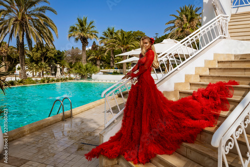 Glamorous Blonde Woman in a Voluminous Red Tiered Gown Posing Dynamically on Marble Stairs by a Luxurious Tropical Resort Pool with Palm Trees and a Bright Blue Sky