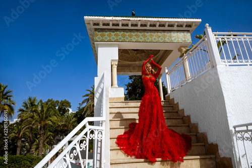 Glamorous Blonde Woman in a Voluminous Red Tiered Gown Posing Dynamically on Marble Stairs by a Luxurious Tropical Resort Pool with Palm Trees and a Bright Blue Sky