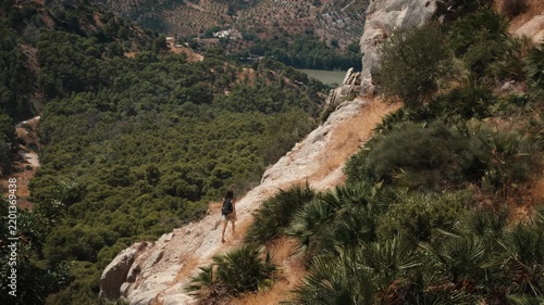 A lone woman with a backpack walking a narrow rocky cliffside path high above a dense forest and reservoir in El Chorro Andalusia Spain under bright natural daylight
