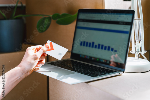 Фотография A person holds a credit card while analyzing financial charts on a laptop screen near moving boxes