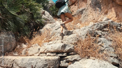 A solo female traveler climbing a steep rocky path with historic ancient stone steps surrounded by dry grass and fan palms under bright sunlight in El Chorro Andalusia Spain