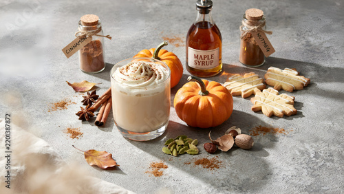 Autumnal pumpkin spice latte served in a glass with maple syrup, cinnamon, nutmeg, and leafshaped cookies on a rustic gray stone background