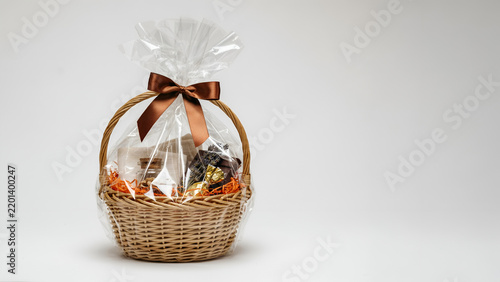 Wicker gift basket filled with assorted treats and wrapped in clear cellophane with a brown ribbon bow isolated on white background