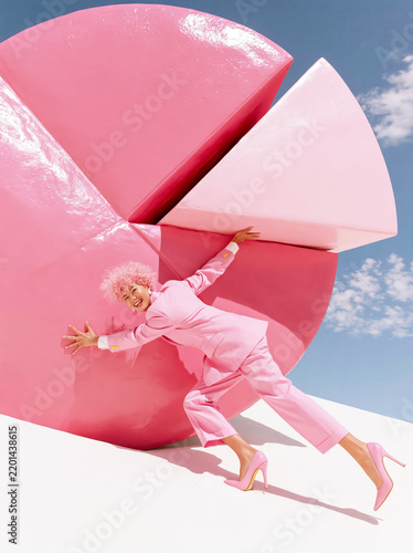 Woman in Pink Suit Pushing Large Pink Geometric Shape Against Blue Sky