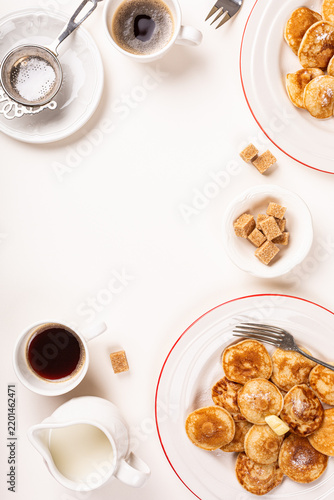 Dutch poffertjes topped with butter sit on red rimmed plates beside coffee, tea, milk, sugar cubes, and utensils, creating a bright overhead breakfast arrangement on a white surface with copy space